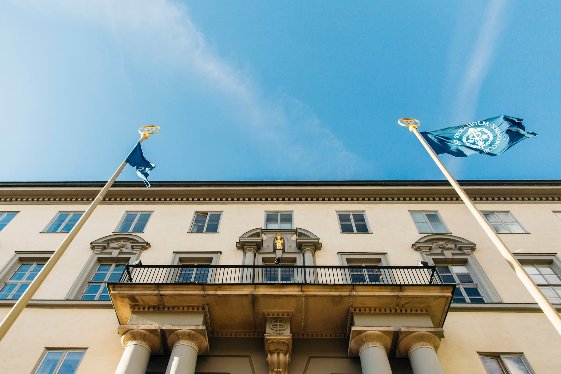 Handelshögskolan i Stockholm vid Sveavägen 65, ljus fasad med balkong, nischstatyett och vajande flaggor.